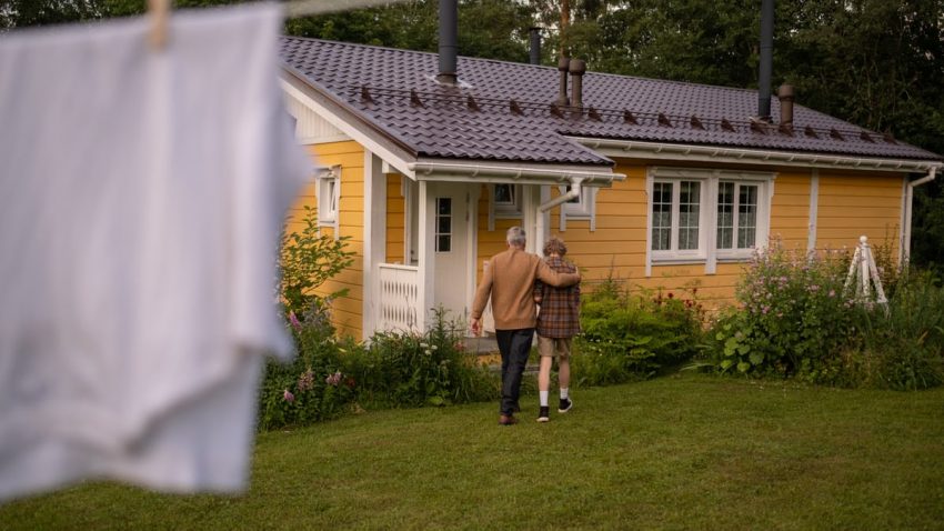 A grandfather and grandson walking towards a quaint yellow cottage in a lush garden setting.
